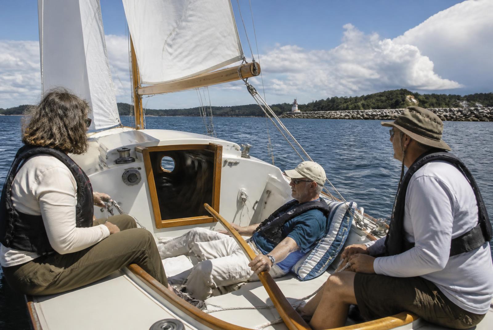 A sailor with a disability seated in the cockpit of an Ensign-class keelboat with the tiller in hand, two crew members alongside for support, sailing near the Marquette breakwater